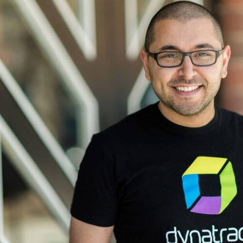 A smiling man wearing glasses and a Dynatrace t-shirt, standing in front of a modern building with geometric patterns